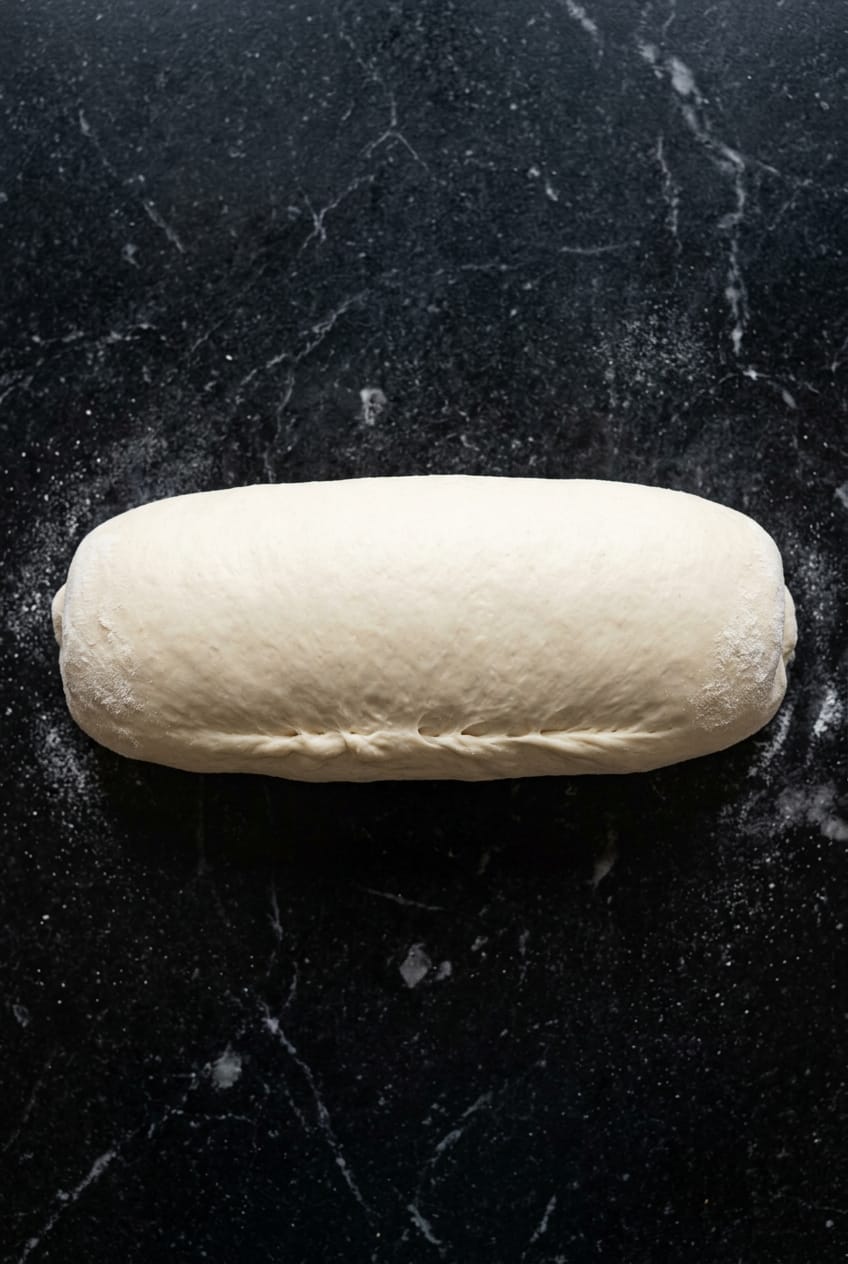 Sandwich bread dough rolled into tight log shape with pinched seam, overhead shot on black marble surface